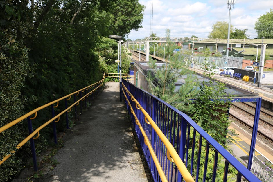 A narrow elevated pathway bordered by bright yellow and blue metal railings winds through a lush, green area adjacent to a railway station. On the left side, dense foliage with various bushes and leaf